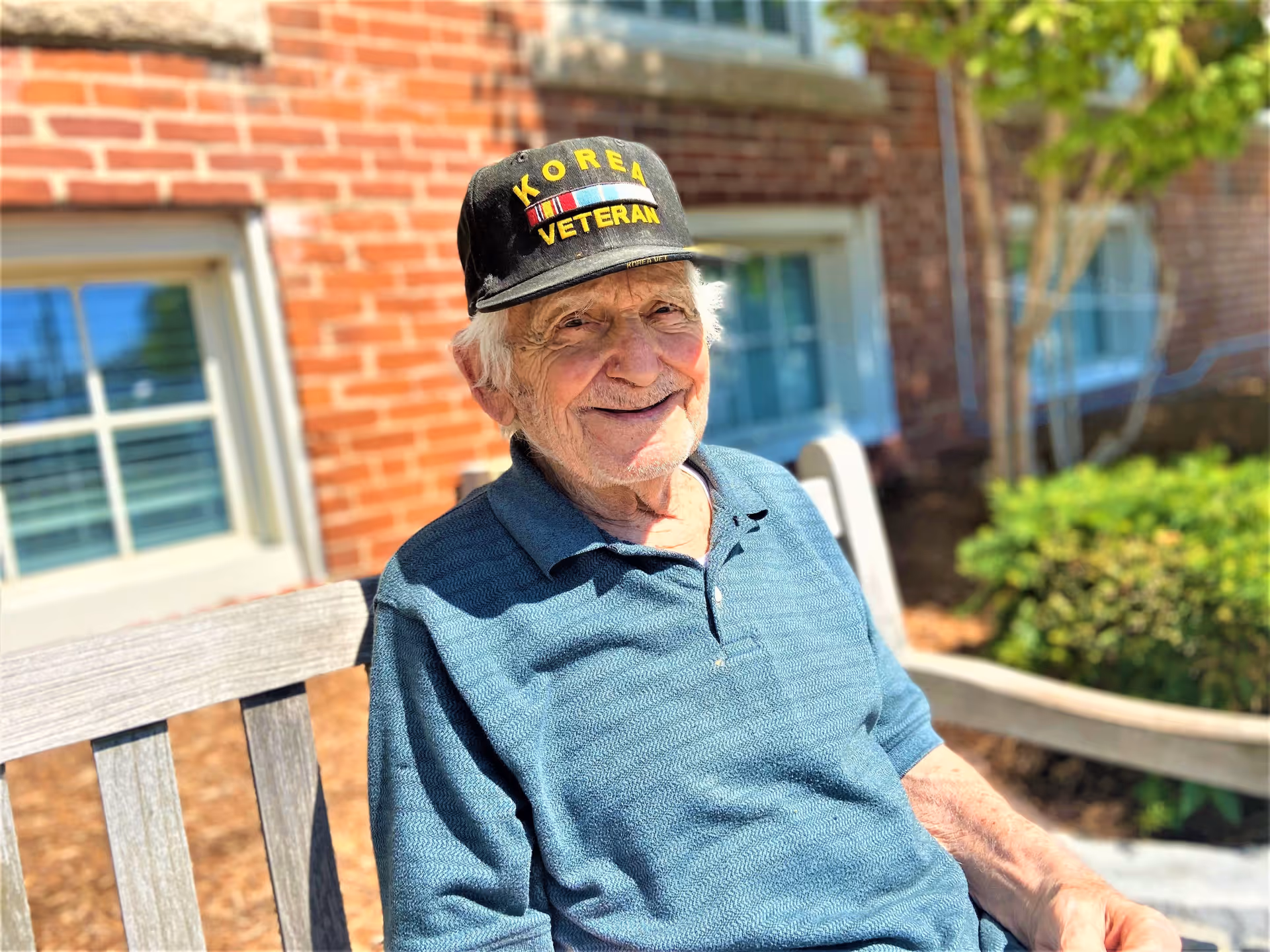 An elderly man wearing a black cap with the words 'Korea Veteran' embroidered on it, sitting on a wooden bench outside a brick building with windows and greenery in the background, smiling at the camera.