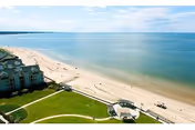Aerial view of a beachfront area with a sandy beach, calm ocean waters, and a grassy lawn adjacent to a building. The sky is partly cloudy with blue tones.