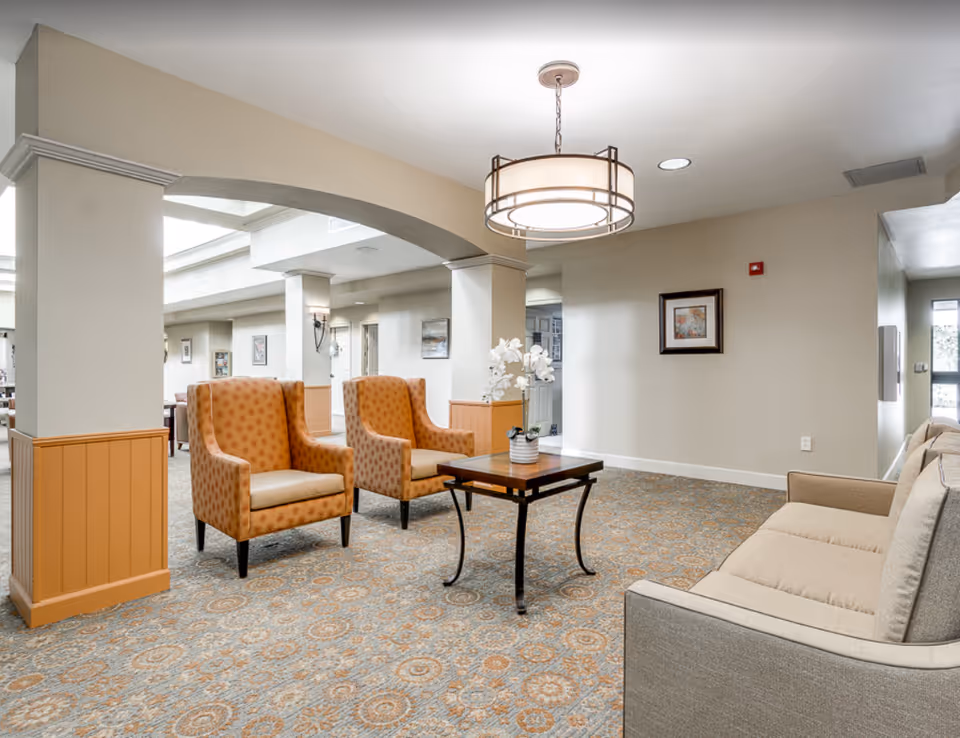 Bright seating area in a senior living facility with two patterned armchairs, a sofa, and a central table under a ceiling light.