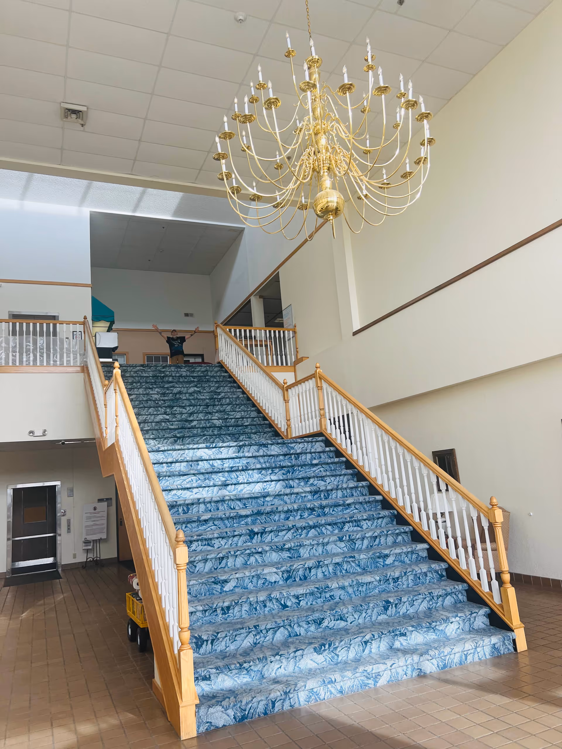 Wide staircase with blue patterned carpet and wooden handrails leading to an upper floor in a building lobby. A large gold chandelier hangs from the ceiling above the staircase. A person stands at the top of the stairs with arms raised. The walls are light-colored and the floor is tiled.