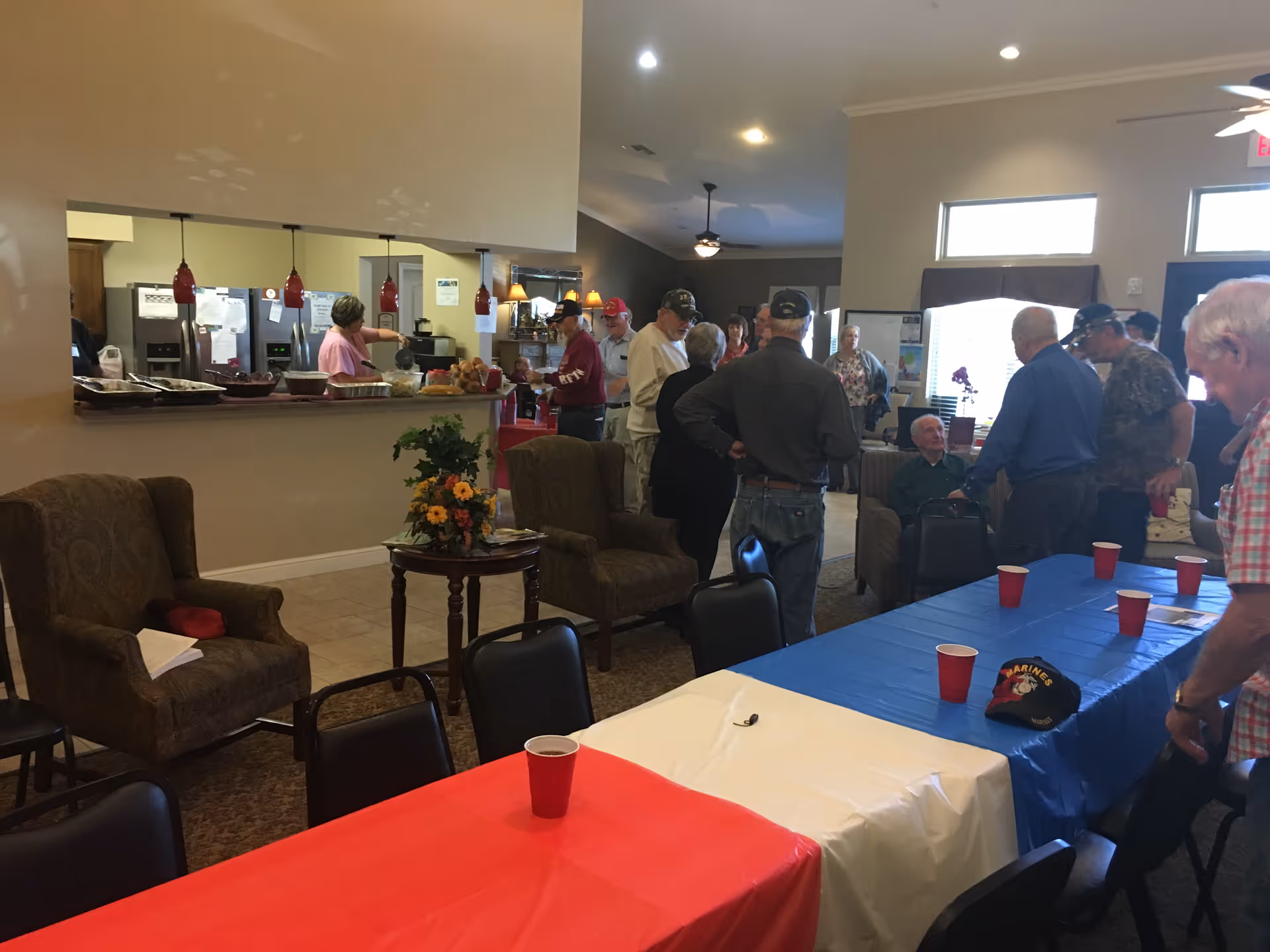 People gather in a senior living common area with long tables covered in red, white, and blue tablecloths, chairs, a serving counter, and armchairs.