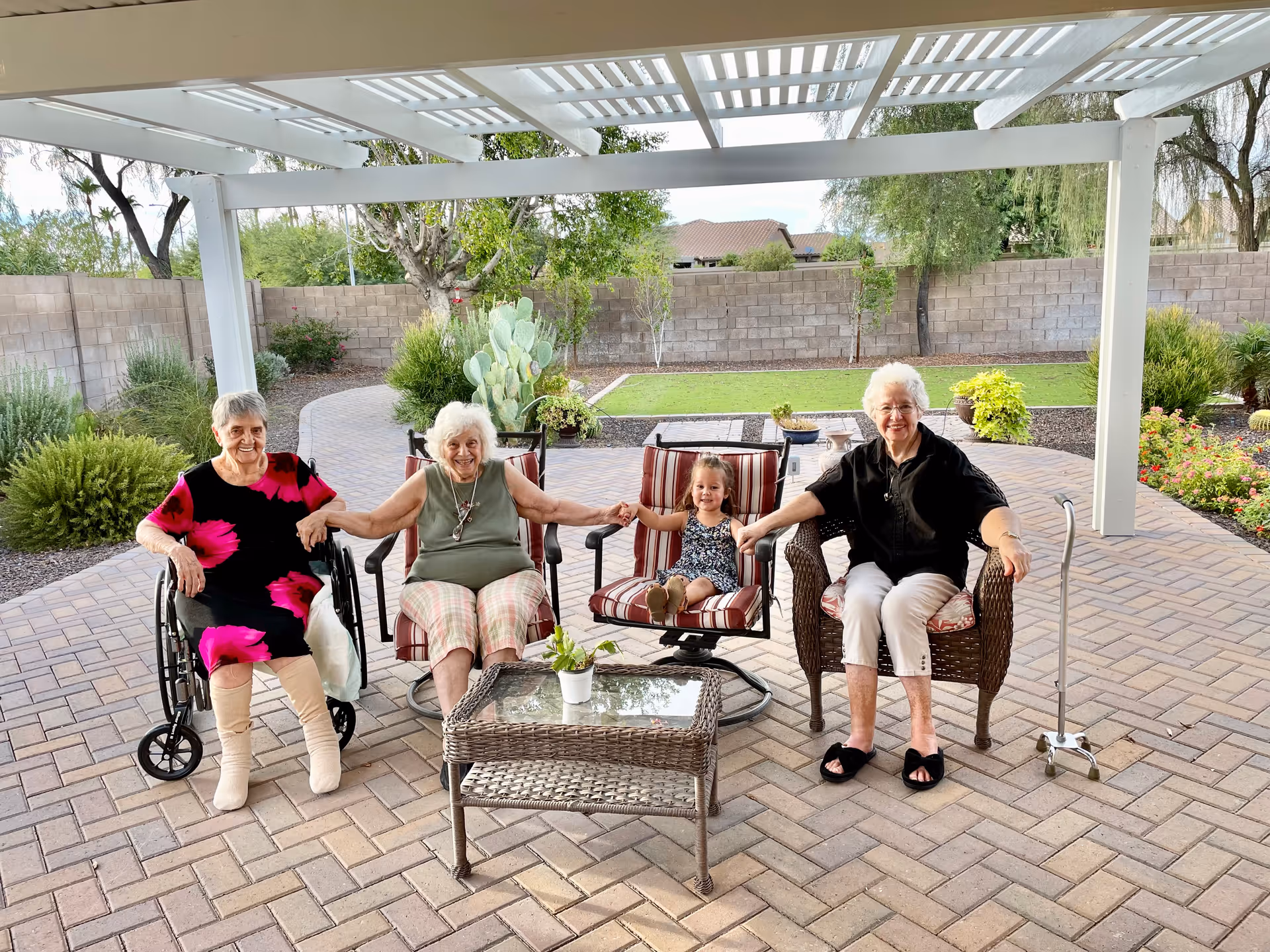 Three elderly women and a young girl sitting outdoors under a white pergola on a paved patio. Two women are seated in chairs, one woman is in a wheelchair, and the young girl is sitting on a cushioned chair between them. They are holding hands and smiling. The background shows a garden with green grass, plants, and a brick wall.