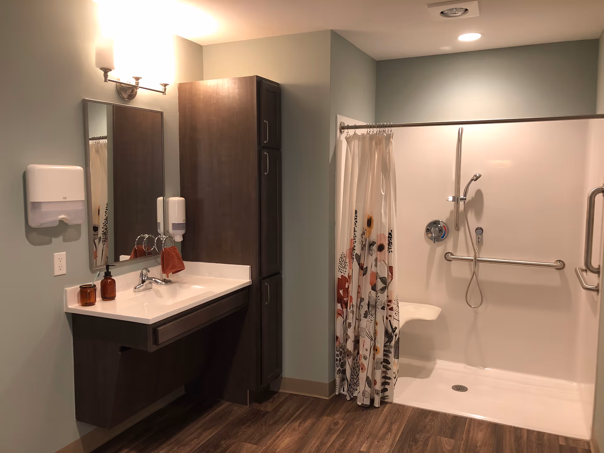 A bathroom with a wall-mounted sink and mirror, a tall wooden cabinet, and a walk-in shower with a floral shower curtain, grab bars, and a fold-down seat. The floor has wood-like flooring and the walls are painted light green.