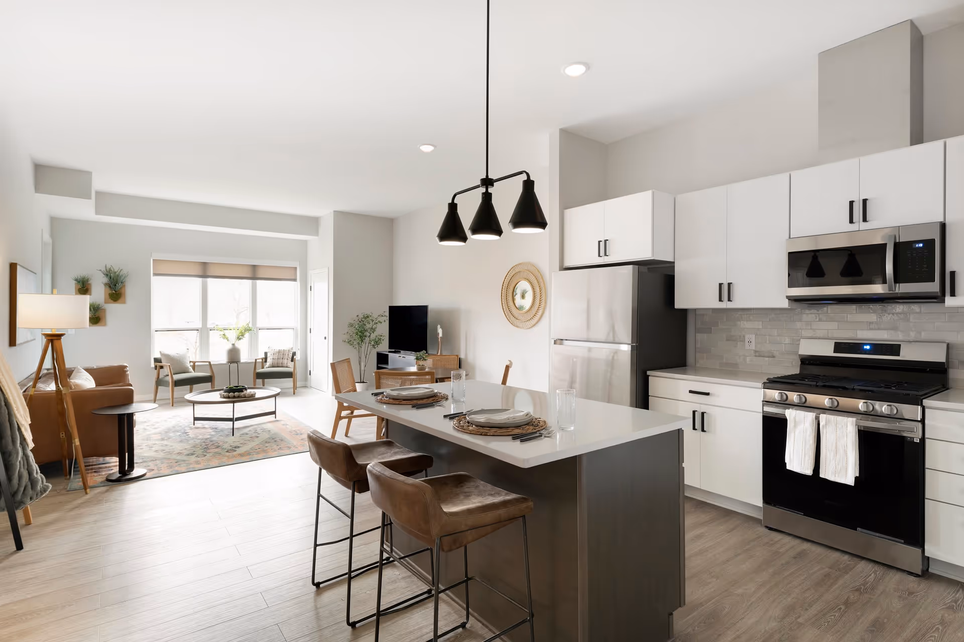 A modern open-concept living space featuring a kitchen with white cabinets, stainless steel appliances, and a kitchen island with two brown bar stools set with plates and glasses. The living area includes a brown leather sofa, two wooden chairs with cushions, a round coffee table on a patterned rug, a floor lamp, and a TV on a stand. Large windows with beige blinds allow natural light to fill the room.
