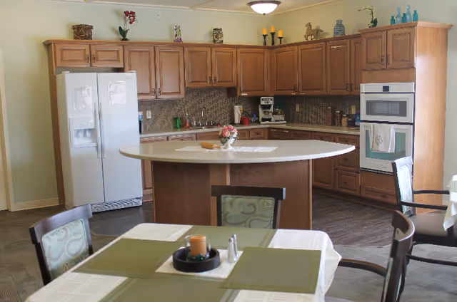 A kitchen area with wooden cabinets, a white refrigerator, a built-in oven, and a central island with a flower vase on top. In the foreground, there is a dining table set with placemats, a candle, and salt and pepper shakers. The kitchen has a tiled backsplash and various decorative items on top of the cabinets.