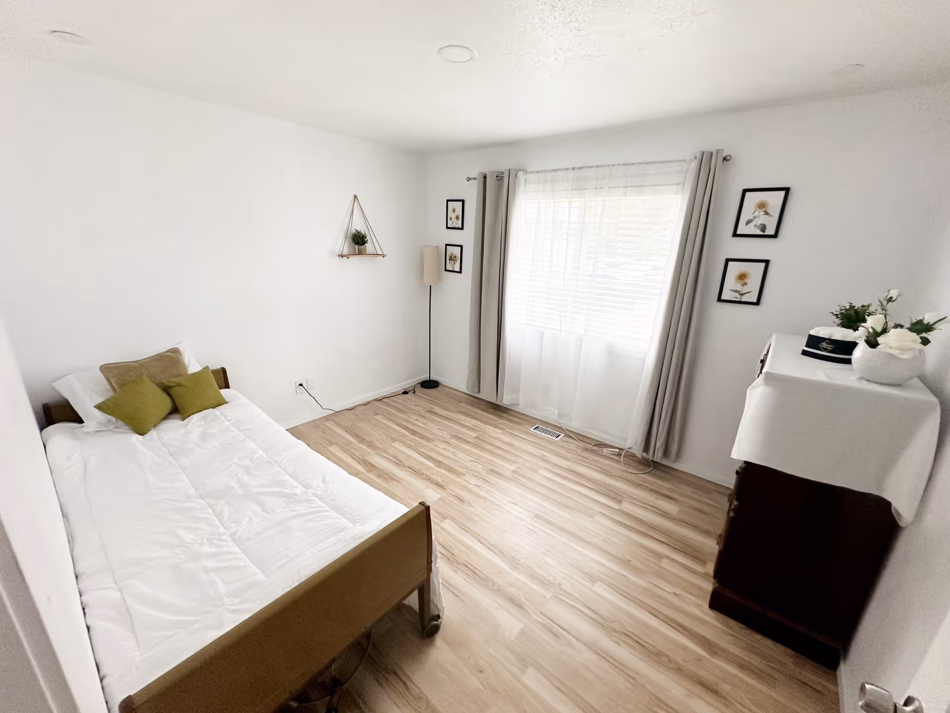 Bright, minimally furnished bedroom with a single bed, window with curtains, a dresser and light wood floors.