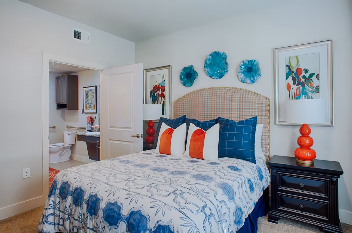 A bedroom with a bed featuring a patterned blue and white bedspread and multiple pillows in blue, white, and orange. The headboard is plaid, and above it are three decorative blue wall hangings. On the right side of the bed is a black nightstand with a red lamp and a framed colorful abstract painting on the wall. An open door reveals a bathroom with a toilet, sink, and cabinets.