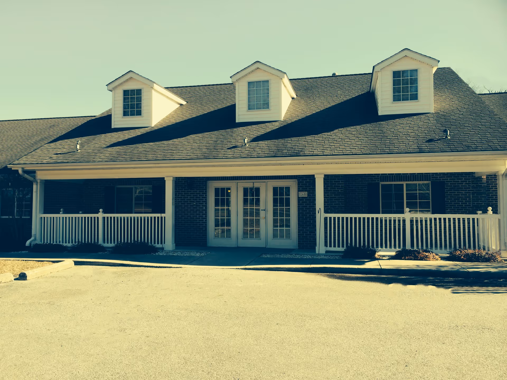 Front exterior view of a single-story brick building with a dark shingled roof featuring three dormer windows. The building has a covered porch with white railings and double glass doors at the entrance. There is a paved parking area in front of the building.