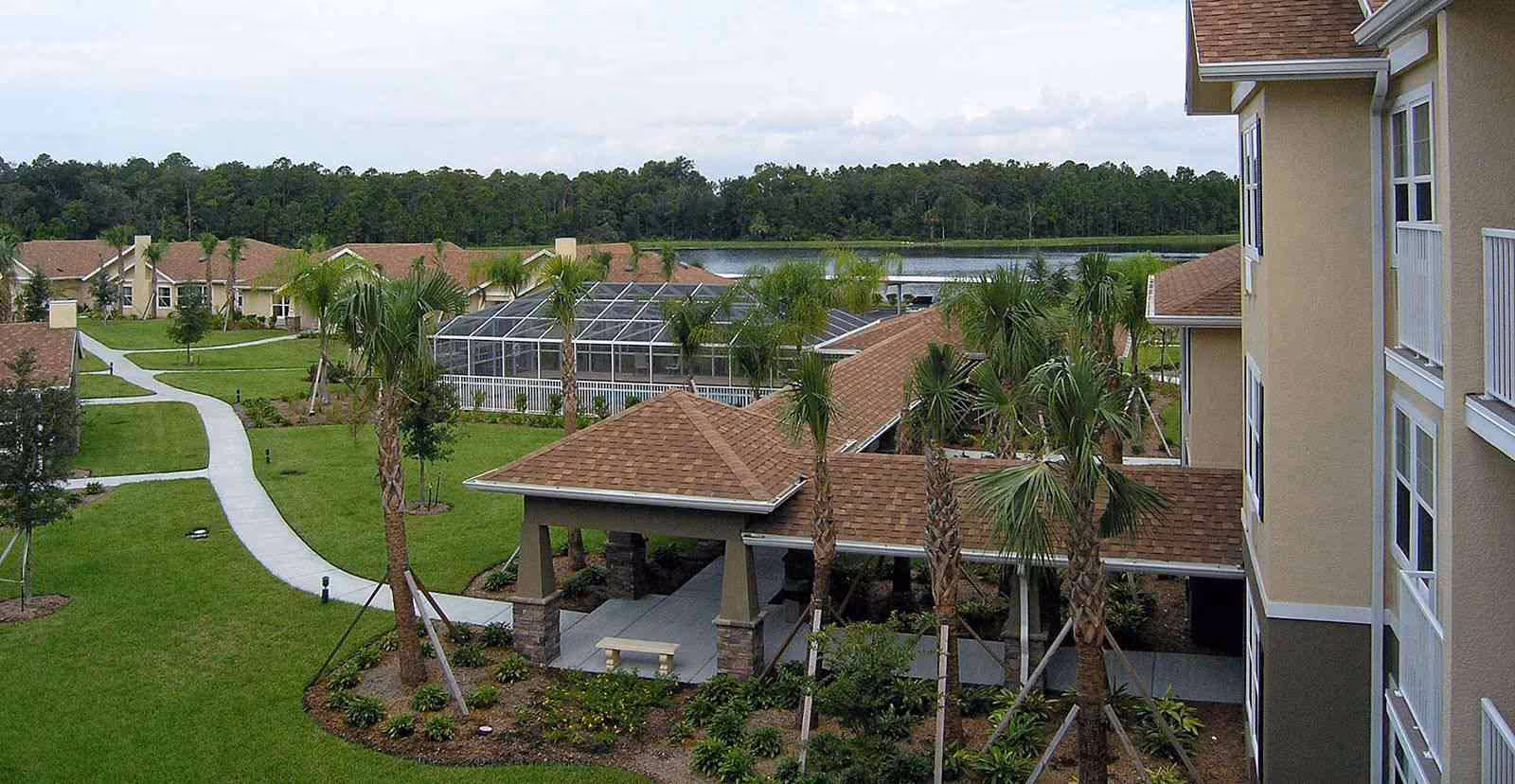 View of a senior living facility with multiple beige buildings featuring brown roofs, surrounded by green lawns, palm trees, and paved walkways. There is a covered outdoor seating area in the foreground and a screened pool enclosure in the middle distance, with a body of water and forested area in the background under a cloudy sky.