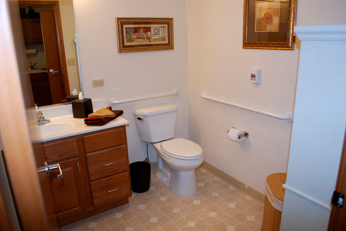 A clean bathroom with a white toilet, wooden vanity with a sink, and a mirror. There are handrails on the walls near the toilet for support. The floor has a beige patterned linoleum, and two framed pictures hang on the walls. A black trash bin is next to the toilet, and a wooden laundry hamper is partially visible.