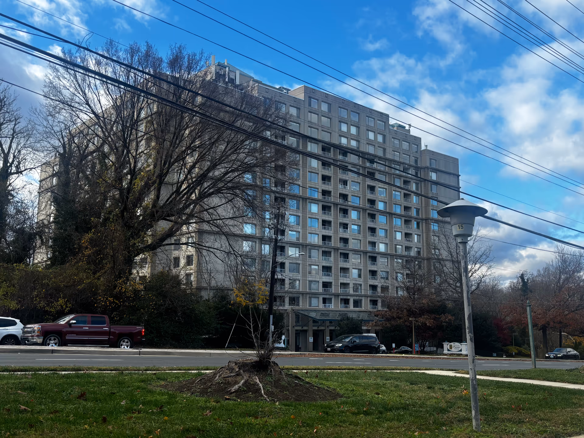 Exterior view of a multi-story residential building with many windows, surrounded by trees with bare branches and some greenery. Several cars are parked along the street in front of the building under a partly cloudy blue sky.