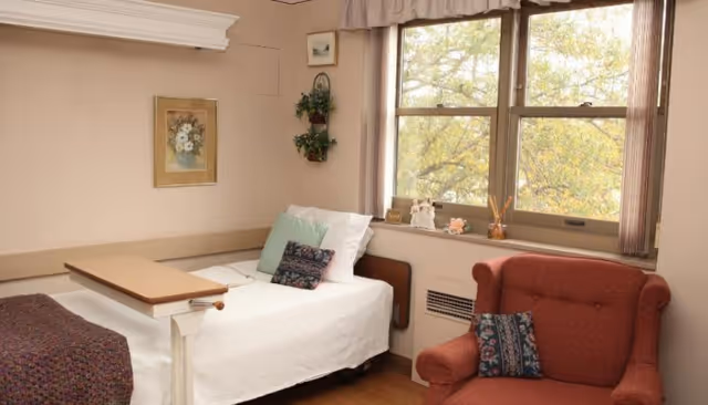A small, cozy room with a single bed covered in white bedding and decorative pillows. Next to the bed is a wooden overbed table. A red upholstered armchair with a patterned pillow sits near a window with beige curtains, through which trees with autumn leaves are visible. The walls are light-colored and decorated with a framed floral picture and a small hanging plant.