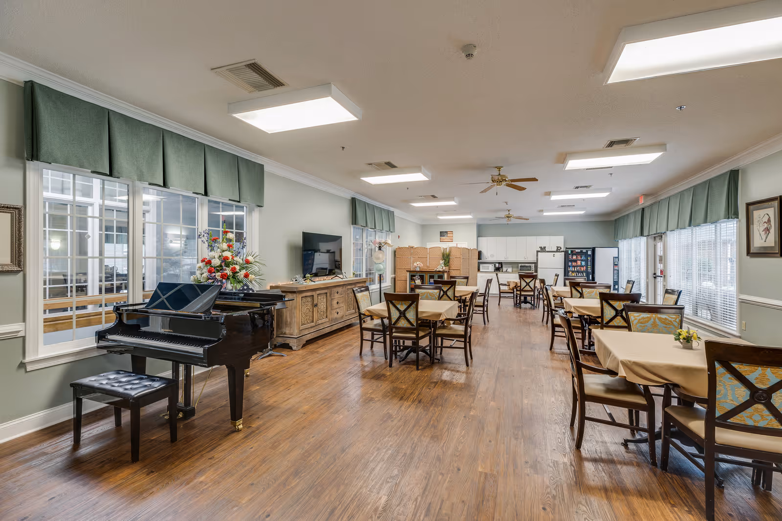 A spacious dining room in an assisted living facility with several tables covered with beige tablecloths and surrounded by chairs. A black grand piano with a matching bench is positioned near large windows with green valances. The room has wooden flooring, ceiling fans, and bright overhead lighting. At the far end, there is a kitchenette area with white cabinets, a refrigerator, a microwave, and a vending machine. The walls are painted light green and decorated with framed artwork.