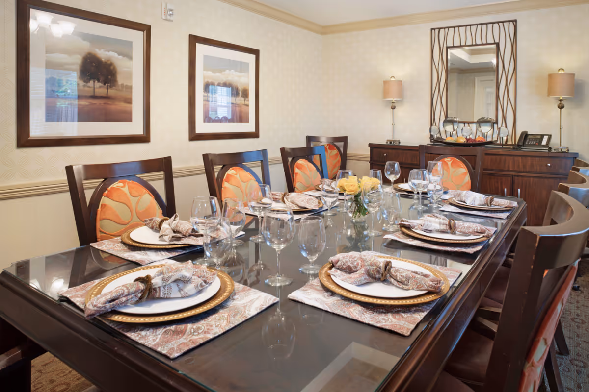 A formal dining room with a glass-top table set for eight people. Each place setting includes a white plate with a gold charger, a patterned cloth napkin with a napkin ring, and multiple wine glasses. The chairs have wooden frames with orange and beige patterned cushions. The room features two framed landscape paintings on the wall, a wooden sideboard with two lamps, a decorative mirror, and a small vase with yellow roses on the table.