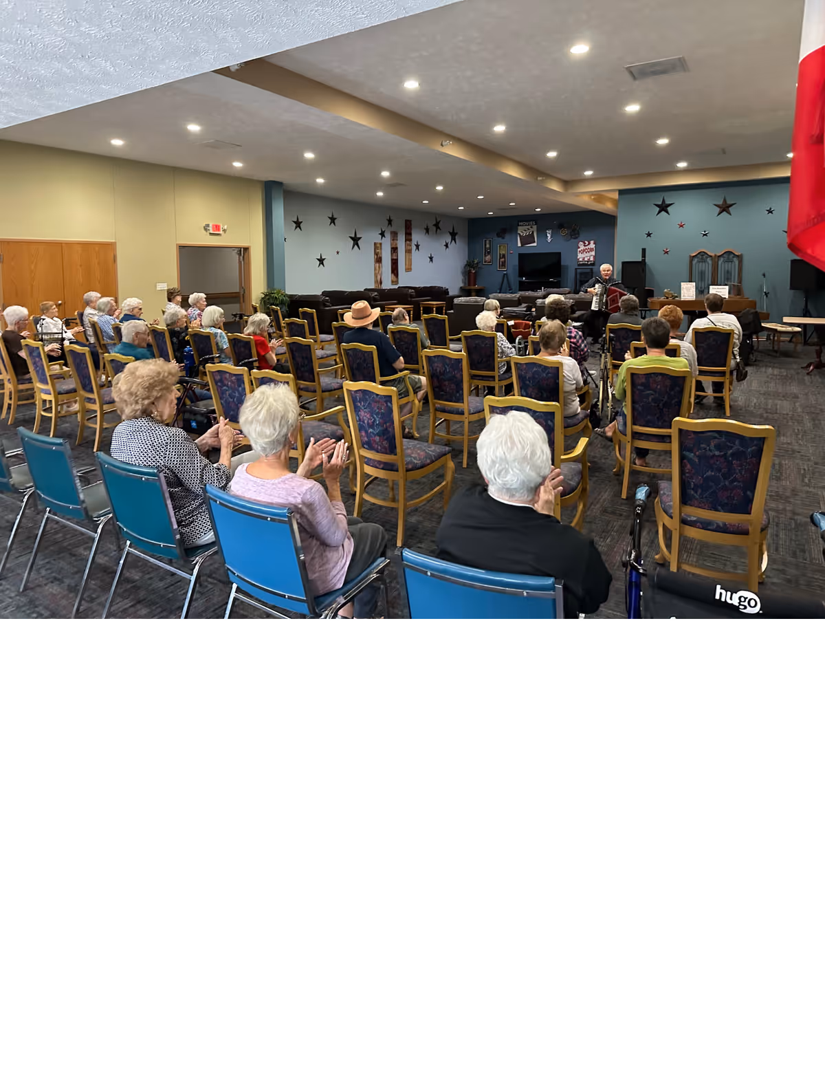 A group of elderly people seated in rows of chairs in a community room, facing a performer playing an accordion. The room has blue walls decorated with star-shaped wall hangings and a television. Some attendees are clapping.