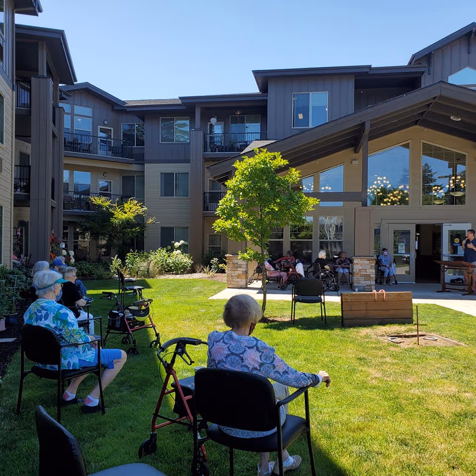 Elderly residents seated on chairs on the lawn of a senior living facility courtyard in front of a multi-story building.