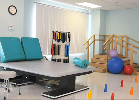 Physical therapy room with a large padded treatment table, a stool, a rack holding various exercise bands, a set of wooden stairs with handrails, and several colorful exercise balls and cones on the floor.