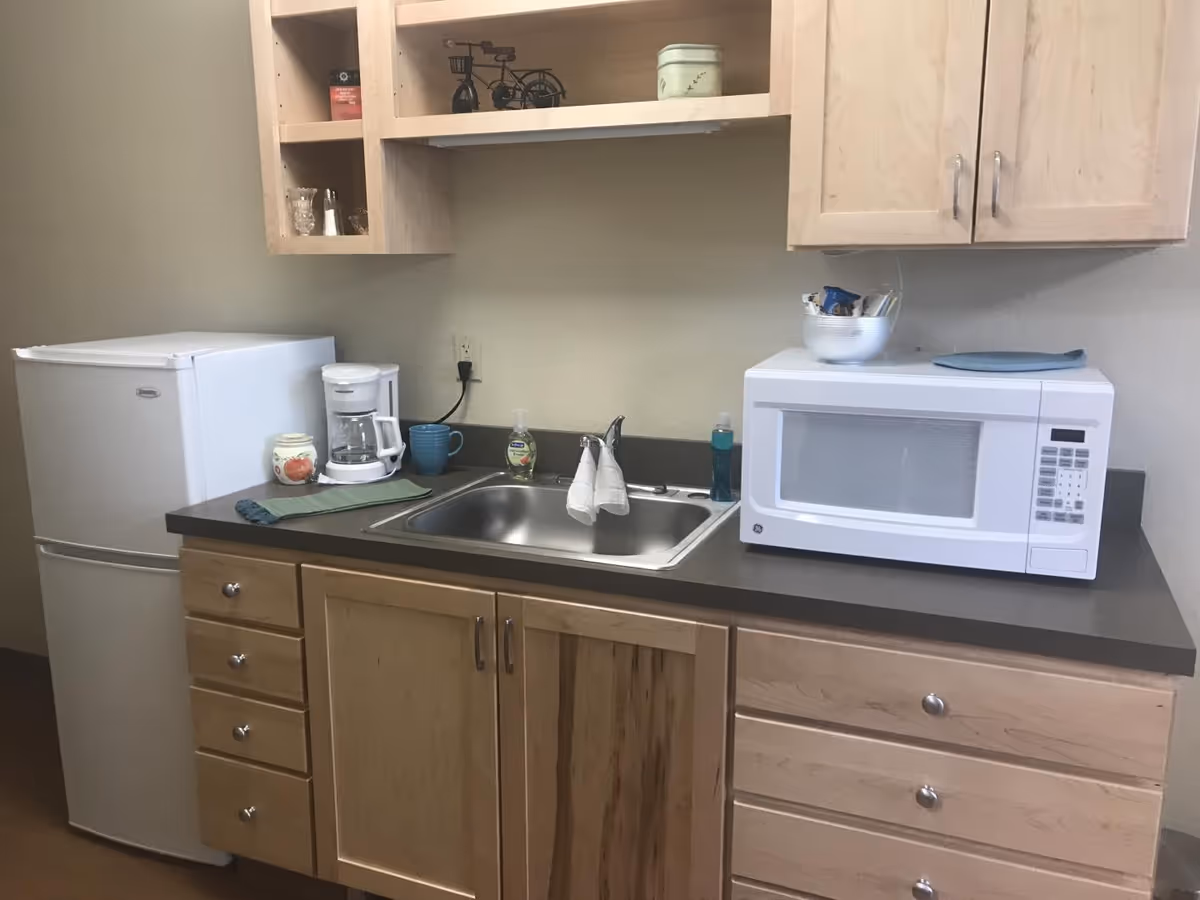 A small kitchen area with light wood cabinets, a white mini refrigerator, a white coffee maker, a blue mug, a stainless steel sink with a faucet, a bottle of hand soap, a white microwave, and a bowl with snacks on the countertop.