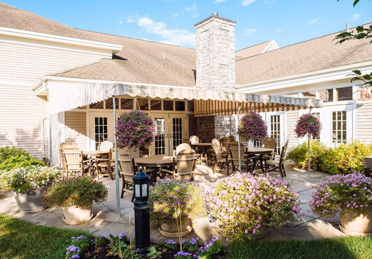 Sunlit outdoor patio with round tables and wooden chairs under a striped awning, surrounded by hanging baskets and potted purple flowers in front of a light-colored building.