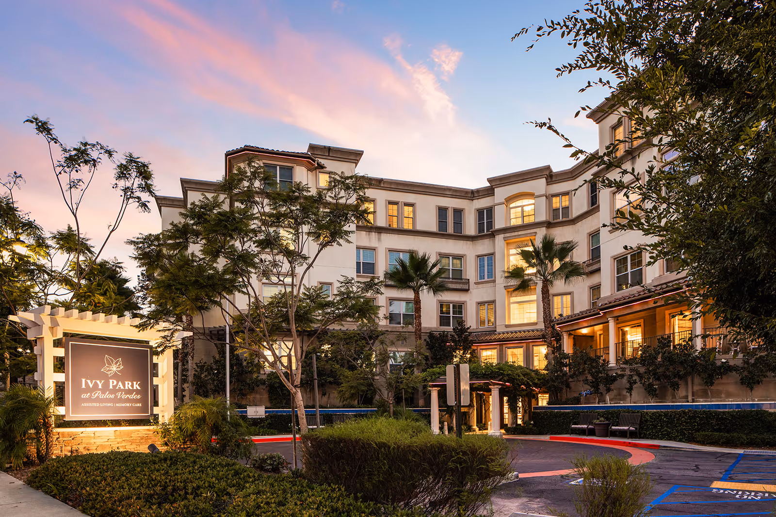 Exterior view of Ivy Park at Palos Verdes senior living facility during sunset, showing a multi-story building with lit windows, palm trees, landscaped bushes, and a sign at the entrance that reads 'Ivy Park at Palos Verdes Assisted Living | Memory Care'.