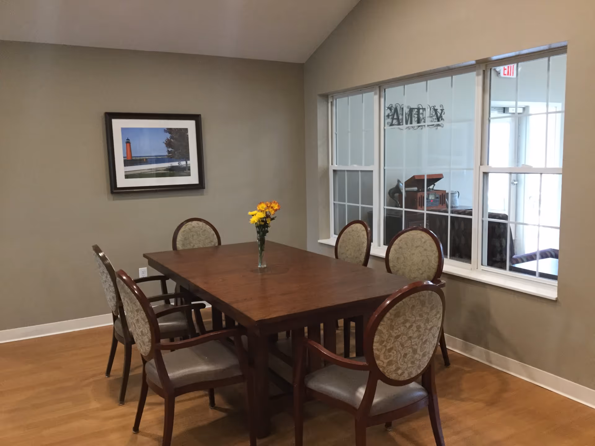 A dining room with a rectangular wooden table surrounded by six upholstered chairs. A vase with yellow and orange flowers is placed in the center of the table. There is a framed picture of a lighthouse on the wall and a large window showing another room with a record player and an exit door.