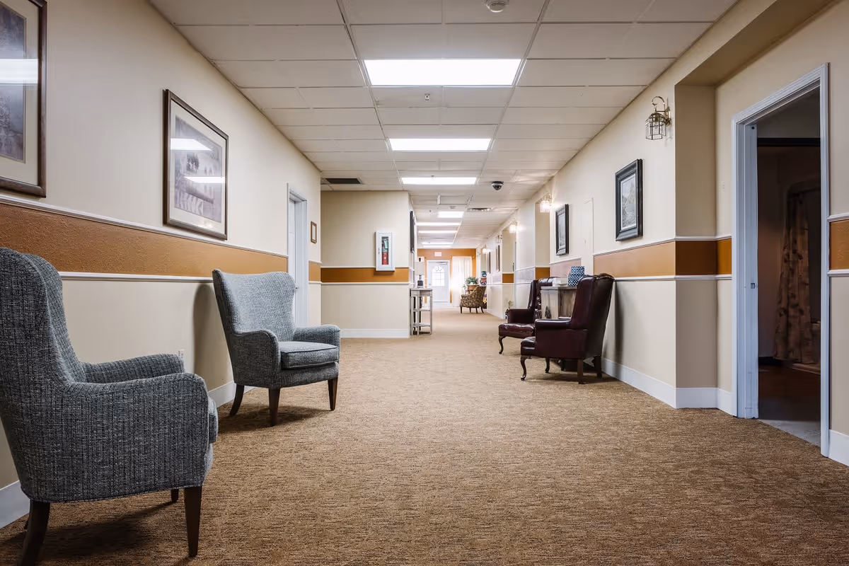 A long, carpeted hallway in a senior living facility with beige and brown walls. The hallway is furnished with several armchairs along the walls and decorated with framed pictures. There are multiple doorways leading to rooms on either side, and the ceiling has recessed lighting panels.