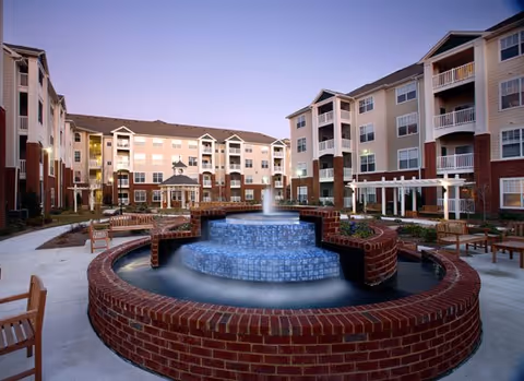 Outdoor courtyard with a tiered circular fountain, benches, and surrounding multi-story apartment buildings at dusk.