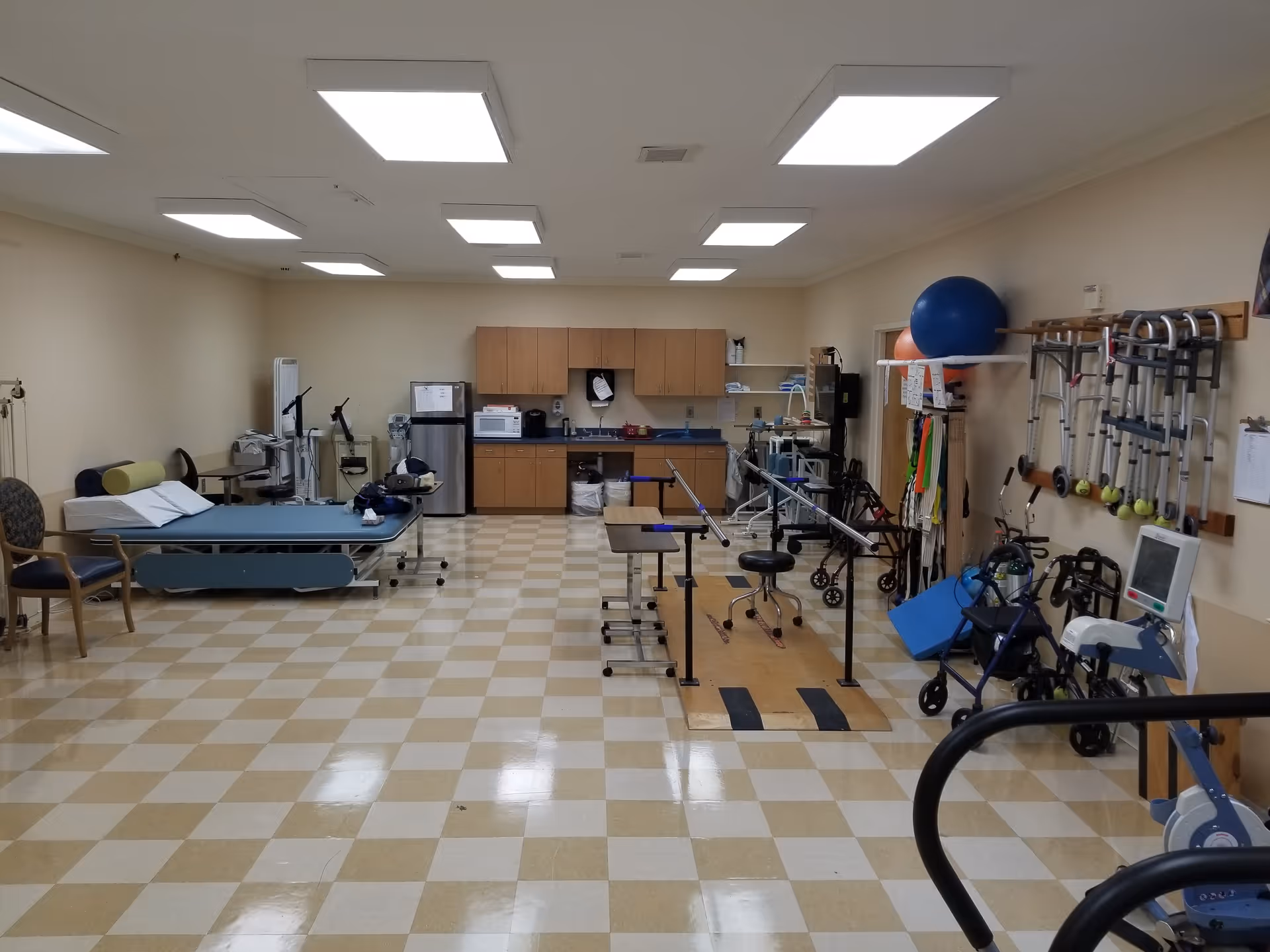 Rehabilitation therapy room with parallel bars, treatment tables, exercise equipment, walkers, and storage cabinets.