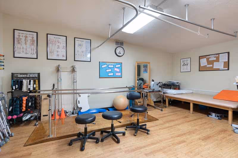 A rehabilitation therapy room with parallel bars for walking exercises, three black rolling stools, various exercise equipment including weights and cones, anatomical charts on the wall, a clock, and a padded therapy table.