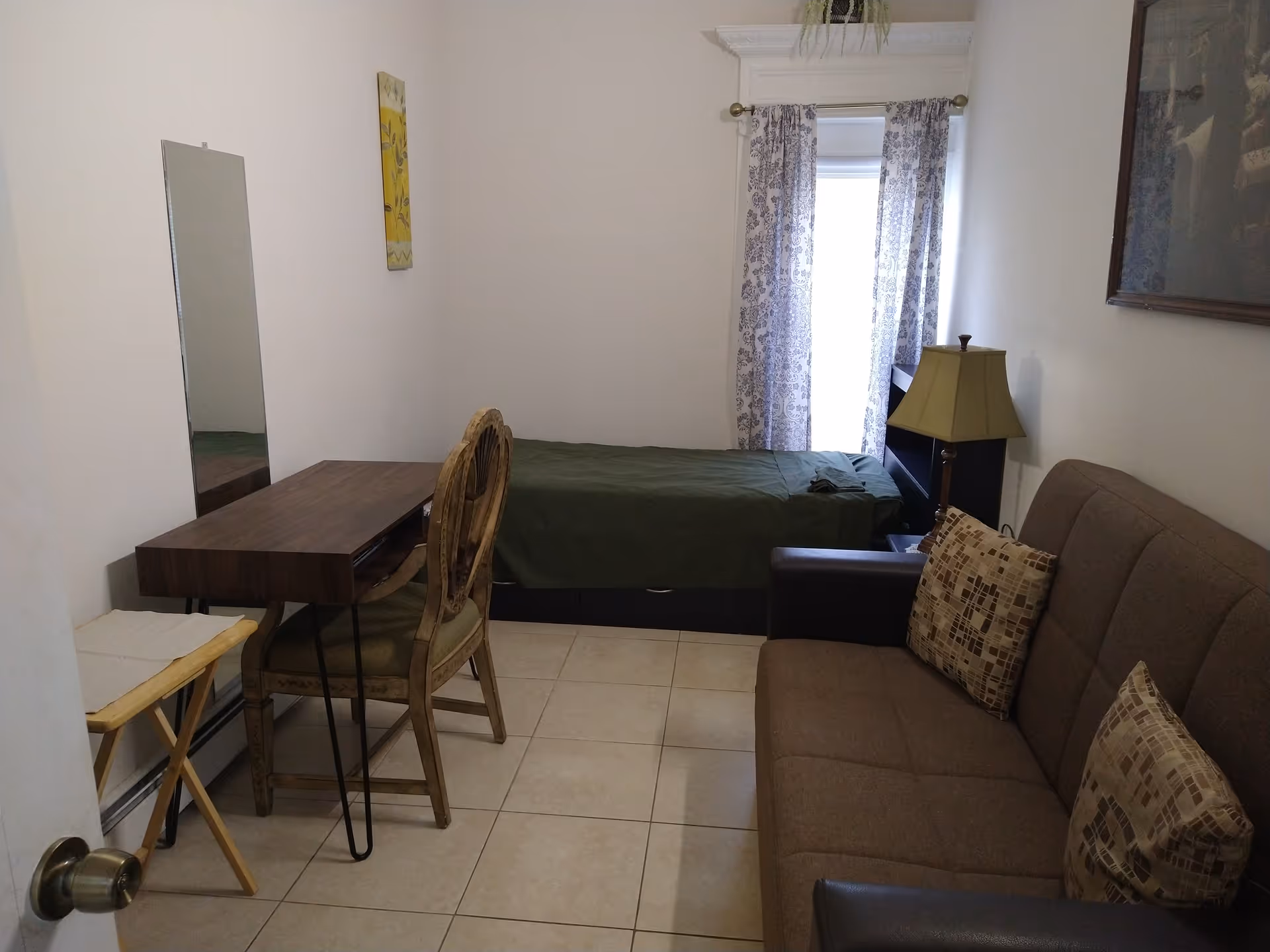 Small furnished room with a single bed against the back wall, a sofa, a desk and chair, and a lamp by a curtained window.