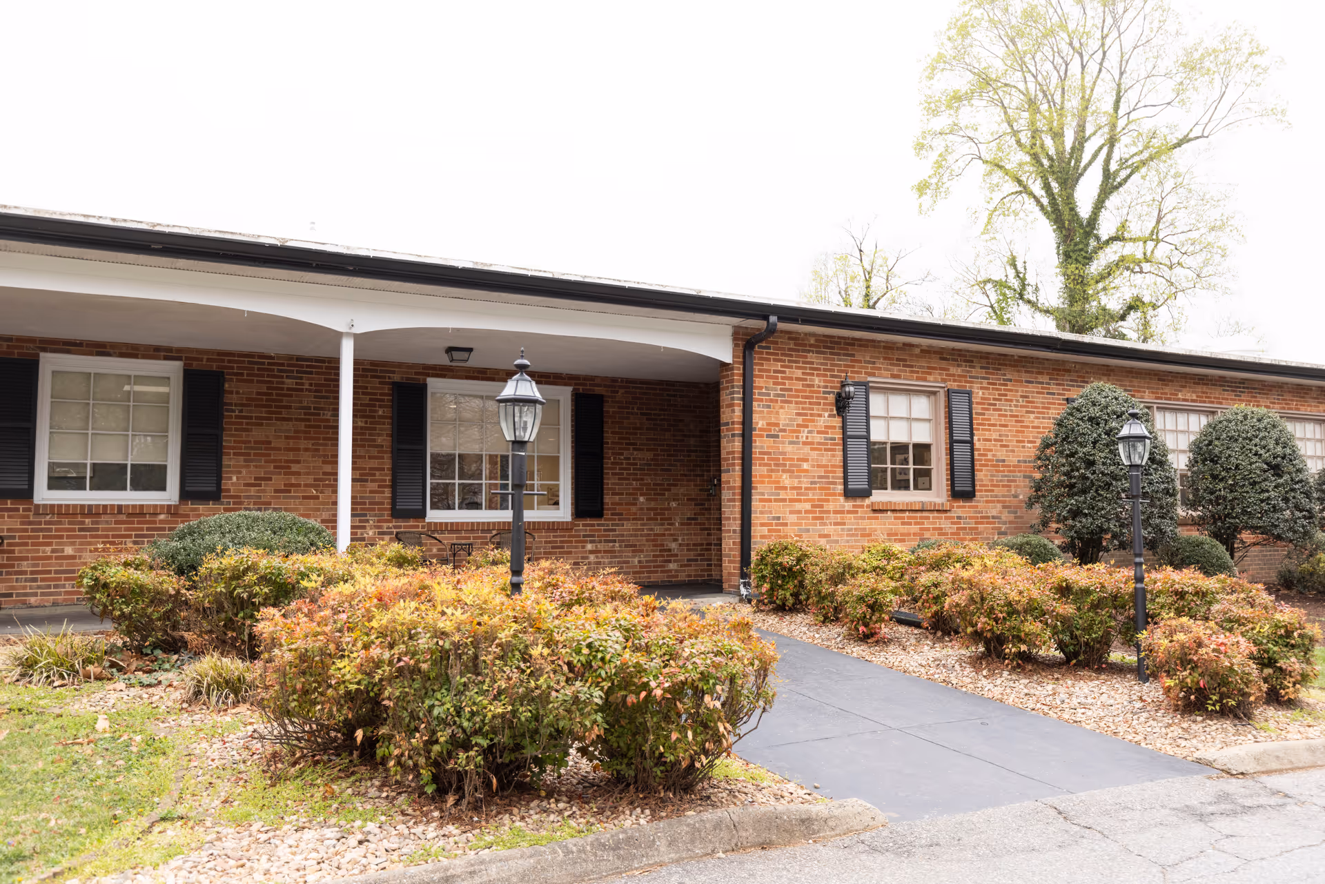 Single-story brick building entrance with a covered porch, walkway, shrubs, and lampposts.