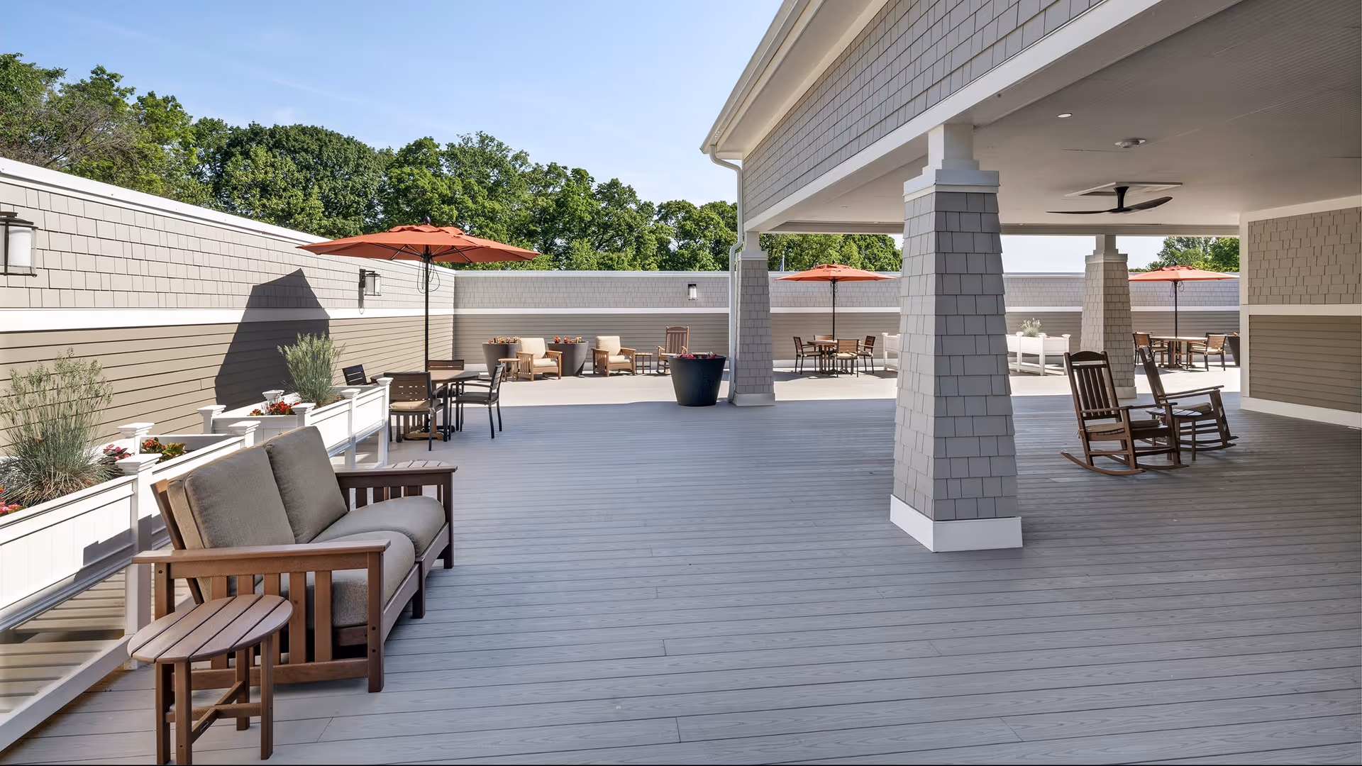 Outdoor patio area with wooden seating including cushioned chairs, rocking chairs, and tables with orange umbrellas. The space is surrounded by a beige and gray wall with planters containing greenery and flowers. Trees are visible in the background under a clear blue sky.