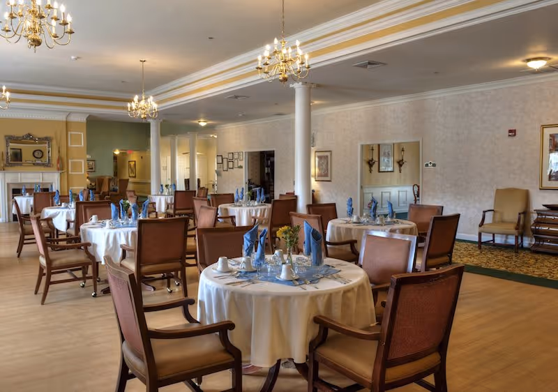 A spacious dining room with round tables covered in white tablecloths, each set with blue folded napkins, cups, and glassware. The room features wooden chairs with armrests, chandeliers hanging from the ceiling, and a fireplace with a mirror above it in the background. The walls are decorated with framed pictures and sconces.