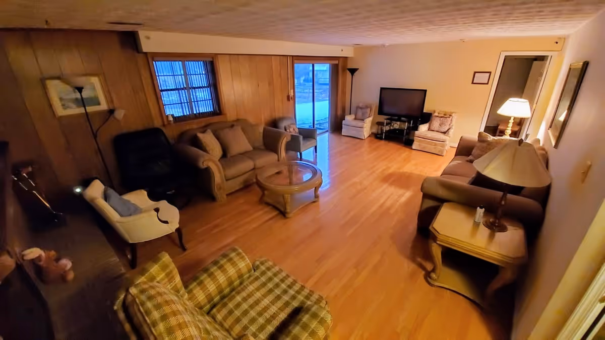 A cozy living room with wooden panel walls and hardwood floors. The room features multiple seating options including a plaid armchair, a beige loveseat, a white armchair, a black recliner, and two cushioned chairs near a flat-screen TV. There is a glass-top coffee table in the center and a wooden side table with a lamp on the right side. A window and a sliding glass door provide natural light, and there are two floor lamps and a framed picture on the wall.