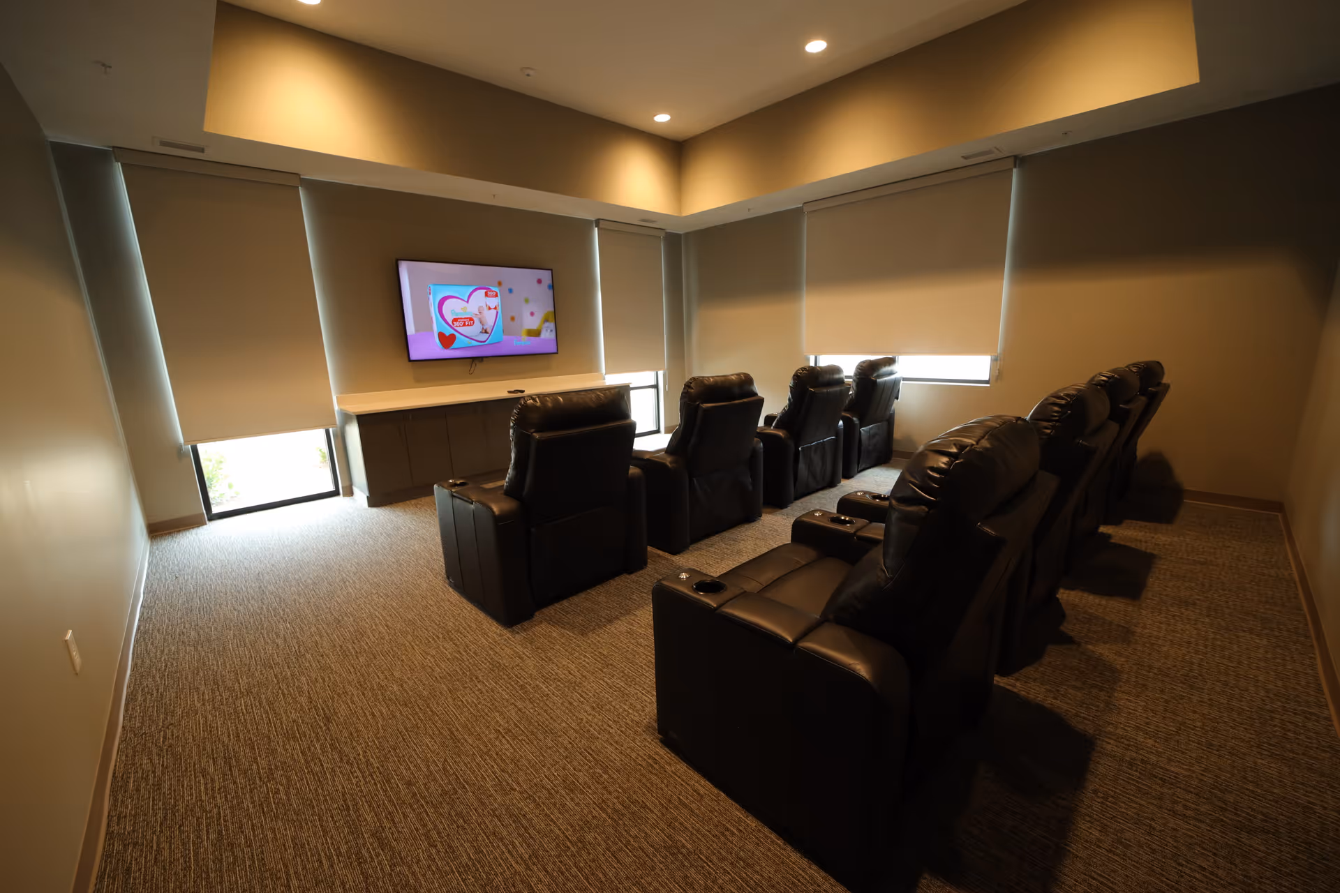 A small theater room with two rows of black leather recliner chairs facing a wall-mounted flat screen TV. The room has beige walls, carpeted floor, and windows with roller shades partially drawn.