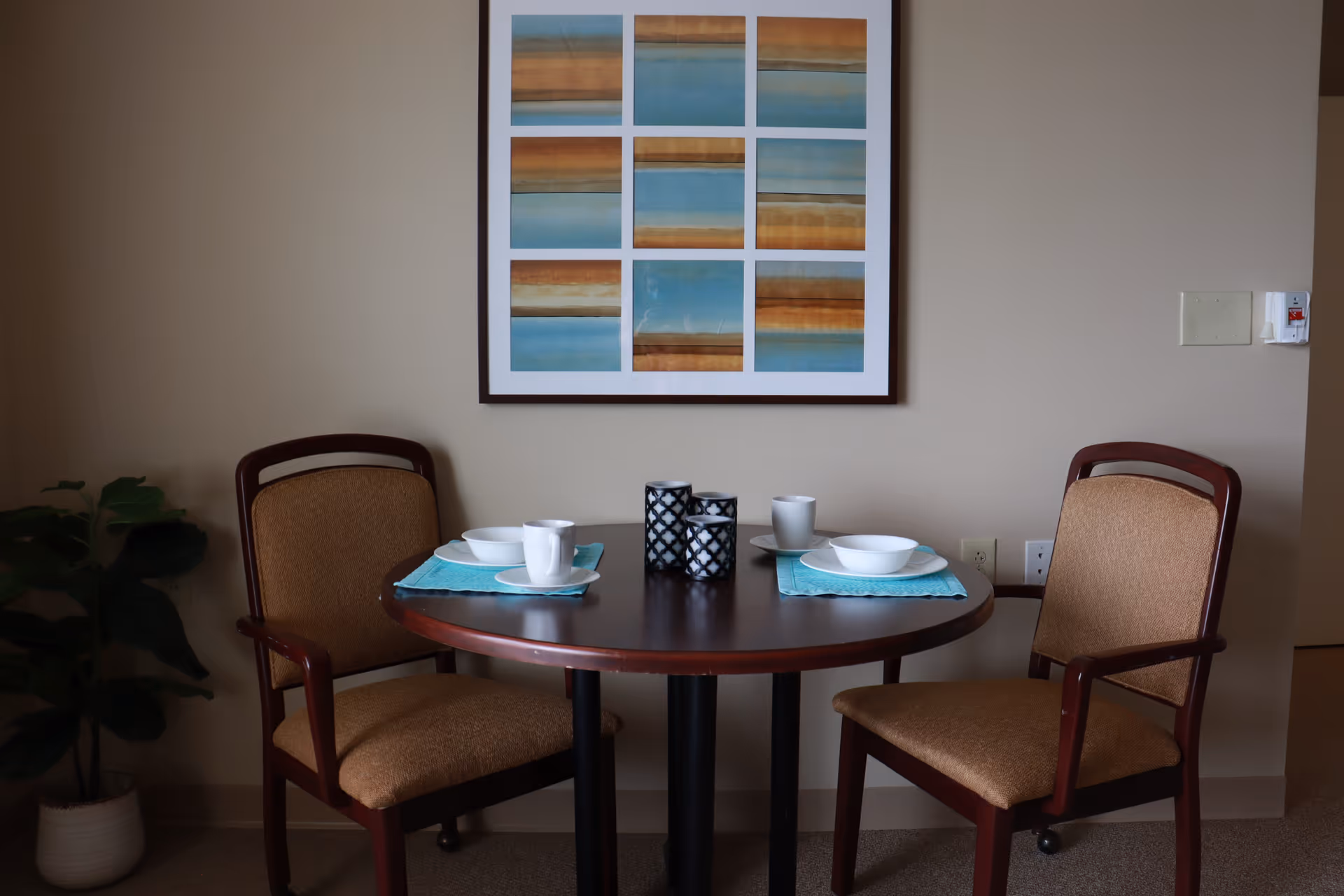 A small round wooden dining table set for two with white bowls, cups, and turquoise placemats. Two cushioned wooden chairs are placed on either side of the table. A framed abstract artwork with blue, brown, and beige tones hangs on the beige wall behind the table. A potted plant is visible to the left of the table.