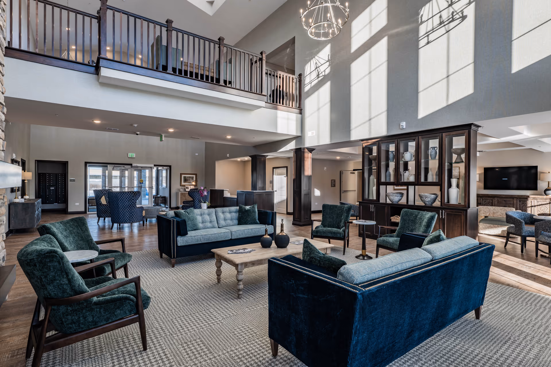 Spacious senior living facility common area with high ceilings and large windows casting light on blue and green upholstered sofas and armchairs arranged around a wooden coffee table on a patterned rug. There is a dark wood display cabinet with decorative vases and bowls, and a TV mounted on the far wall. The area includes a balcony railing above and multiple seating areas with chairs and tables.