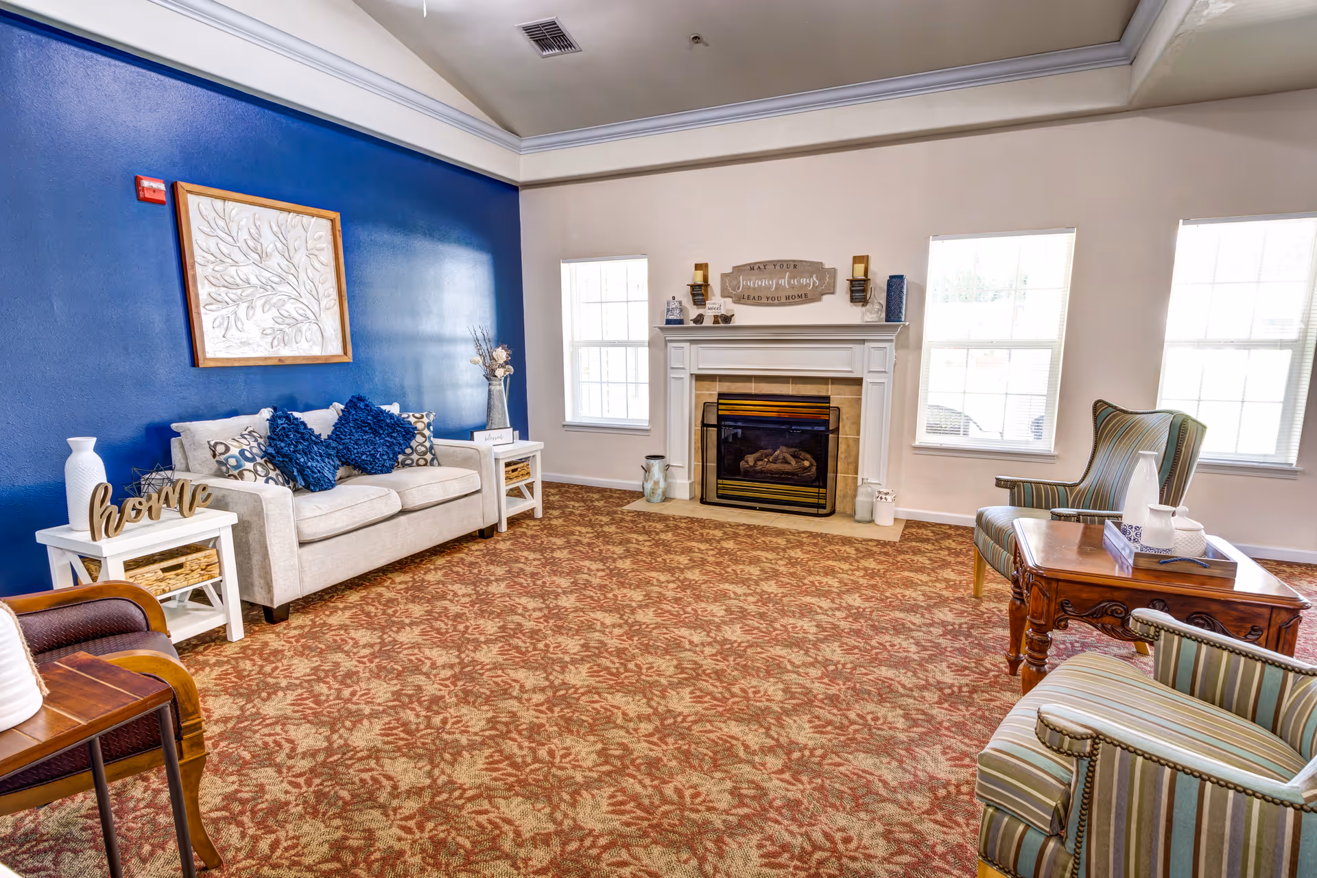 A cozy living room with a beige sofa adorned with blue and patterned cushions against a blue accent wall. There are two white side tables with decorative items, a fireplace with a mantel decorated with candles and a sign, and two striped armchairs with a wooden coffee table in between. The room has three large windows allowing natural light to fill the space and a patterned carpet covering the floor.