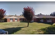Single-story brick building with a green lawn and a few small trees in front under a clear sky, likely a residential or senior living facility.