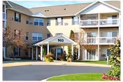 Front entrance of a three-story senior apartment building with a covered porte-cochere, balconies, and landscaping.