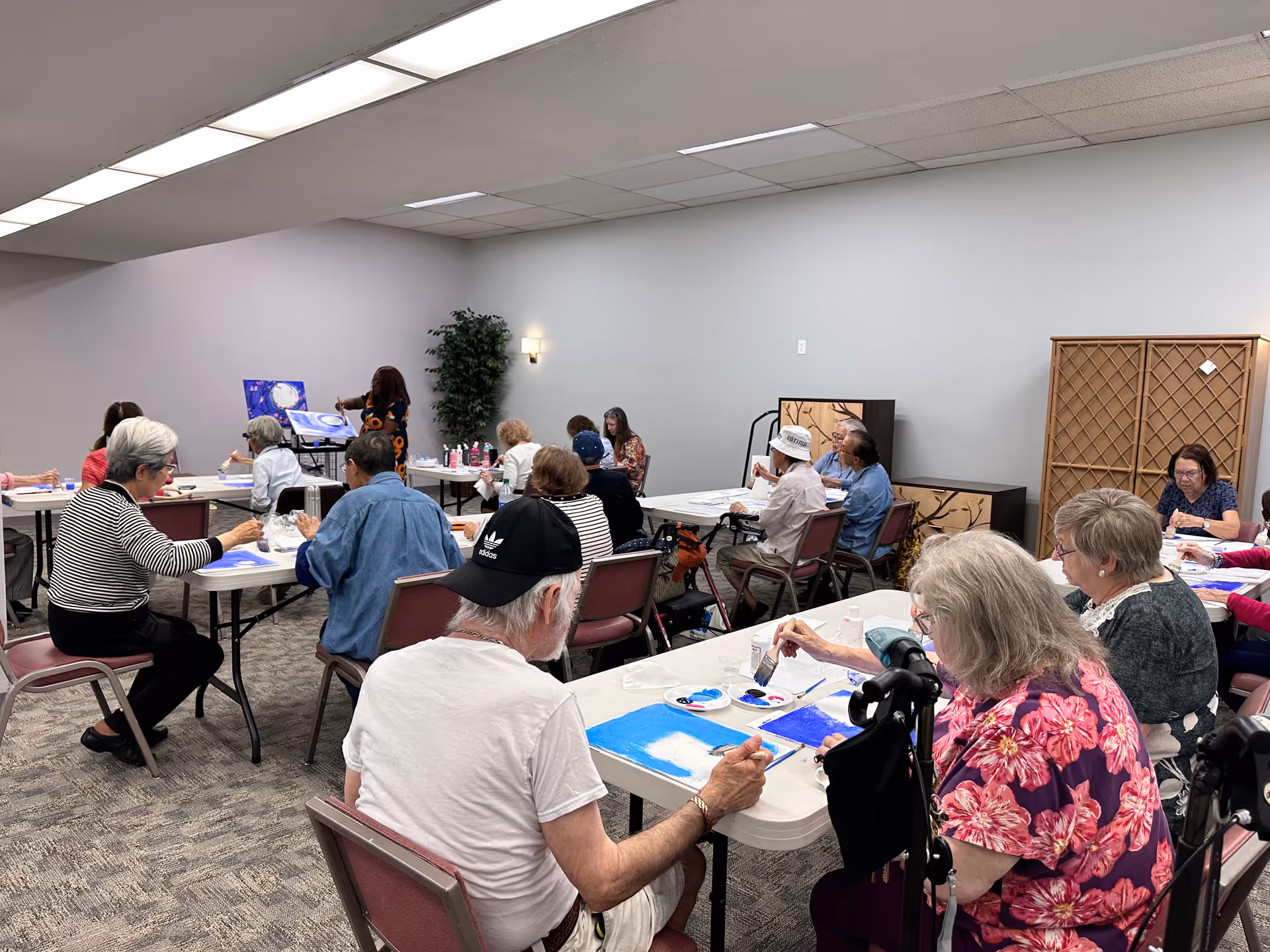 A group of elderly people seated at tables in a room, participating in a painting activity. They are painting on canvases with brushes and blue paint. The room has plain white walls, overhead fluorescent lighting, and a carpeted floor. A woman stands at the front near an easel demonstrating painting techniques.
