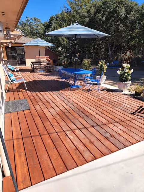 Outdoor wooden deck area with two blue and white striped umbrellas providing shade over a blue metal table and chairs set and a wooden picnic table with benches. There are white metal chairs with blue cushions along the side of the building. Potted plants and trees are visible in the background under a clear blue sky.