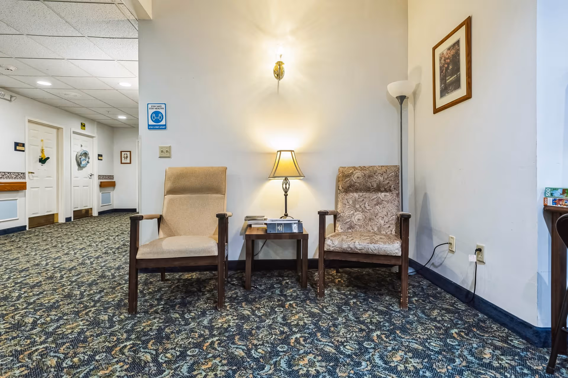 Two armchairs with wooden frames and different upholstery patterns are placed on either side of a small wooden table with a lamp and some books on it. The setting is in a hallway with patterned carpet, white walls, a framed picture, and a floor lamp in the corner. Doors and a hallway are visible in the background.