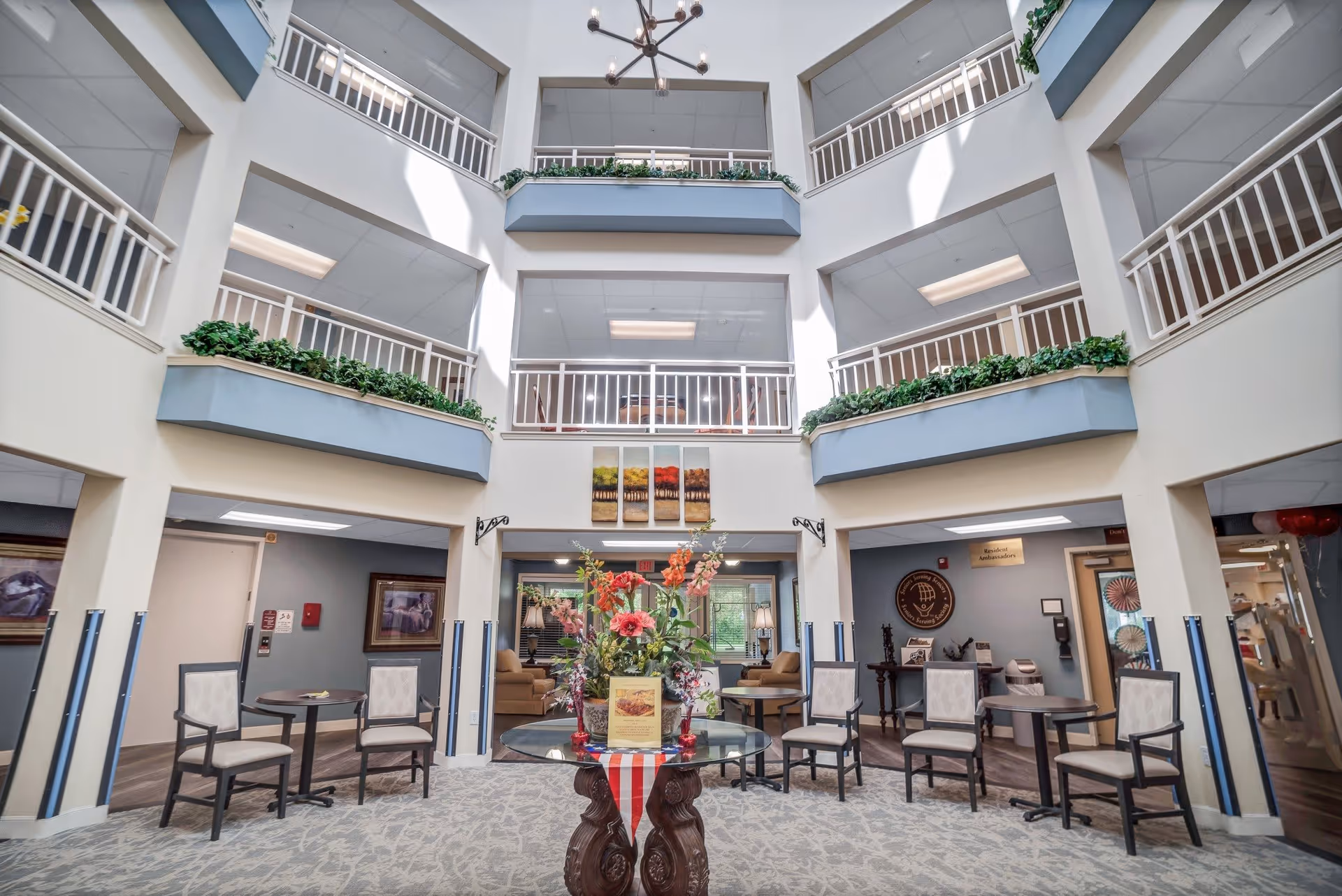 Interior view of a multi-story senior living facility atrium with balconies on upper floors, a central round table with a floral arrangement, several chairs and small tables arranged around the space, and artwork on the walls.