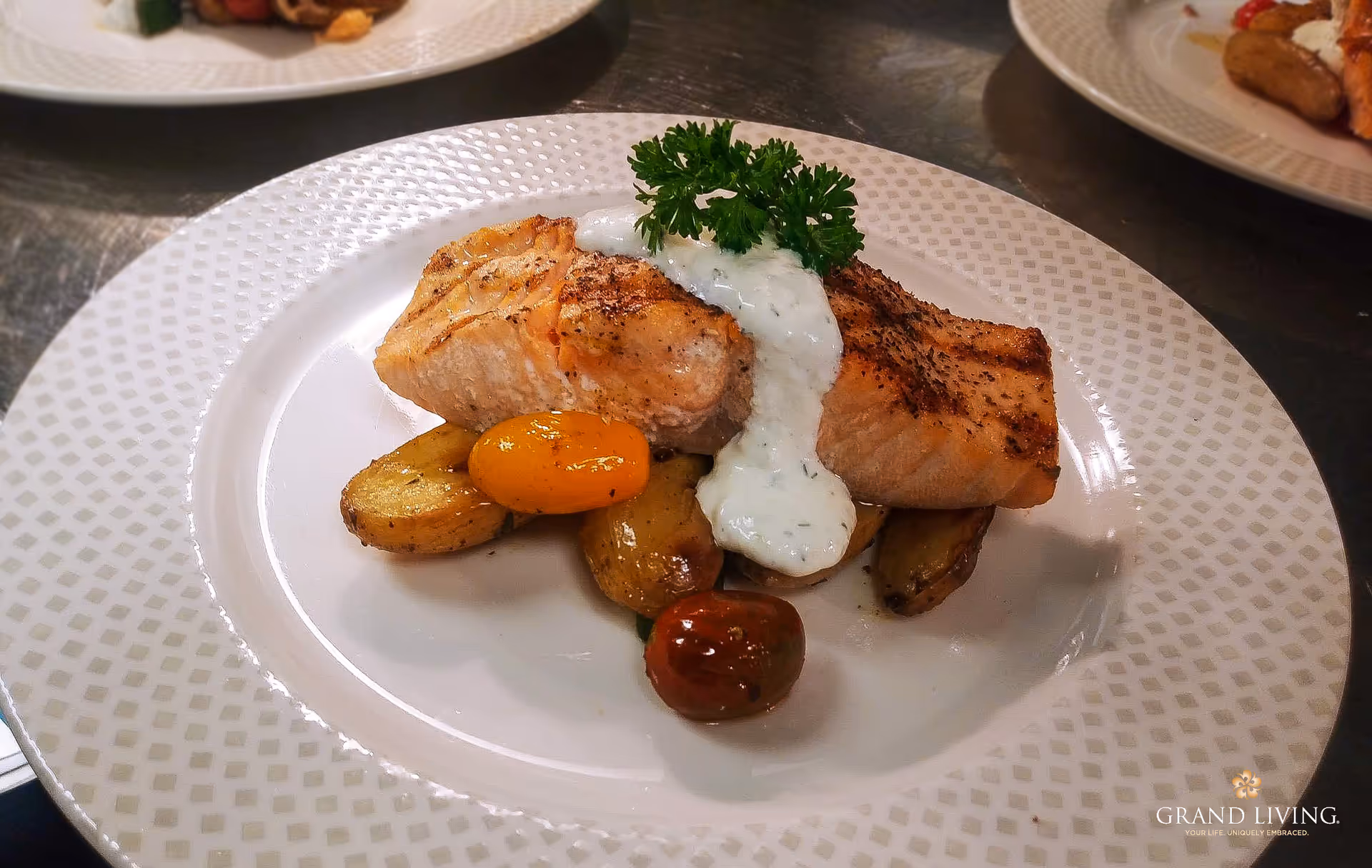 Grilled salmon fillet on roasted potatoes and cherry tomatoes topped with a creamy herb sauce and parsley on a white patterned plate.