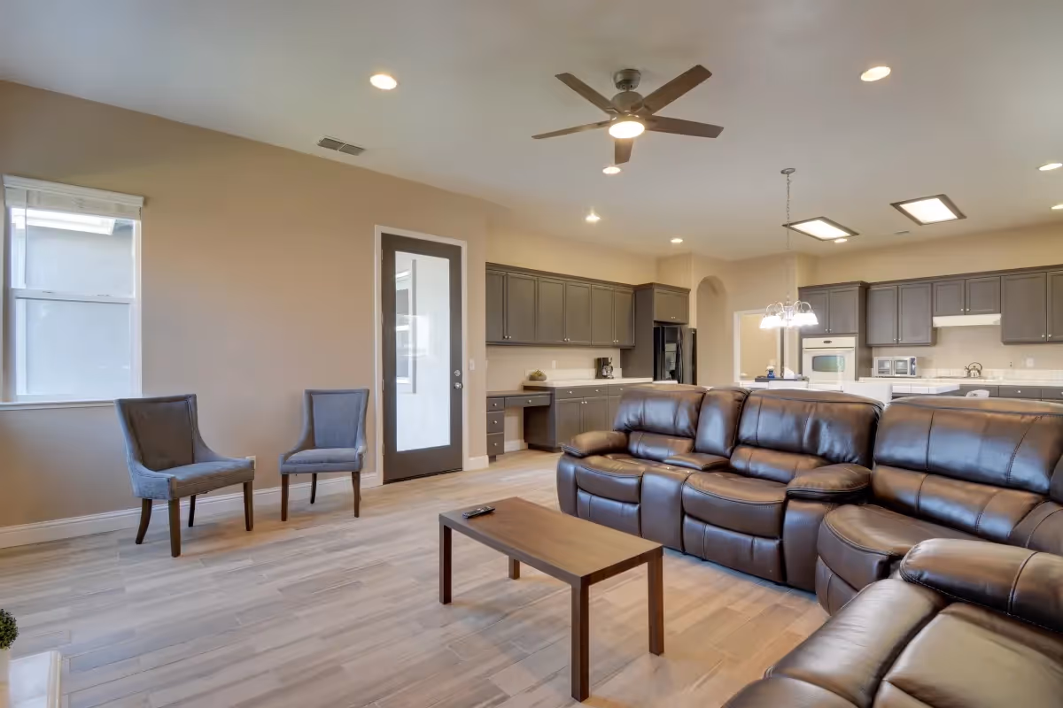 A spacious, modern living area with a large brown leather sectional sofa, a wooden coffee table, two gray upholstered chairs near a window, and an open kitchen with gray cabinets and white countertops in the background. The room has light wood flooring, recessed lighting, a ceiling fan, and a glass door.