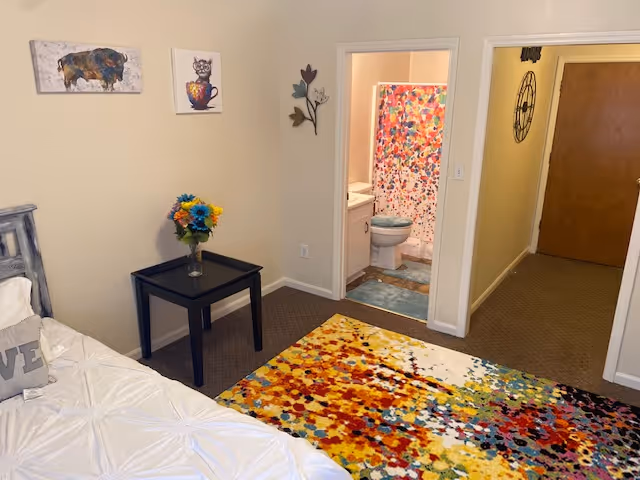 A cozy bedroom with a white bedspread and a decorative pillow on the bed. A small black side table holds a vase with colorful flowers. The room features a vibrant multicolored rug on the floor. On the wall above the side table are two pieces of artwork: one of a buffalo and another of a cat in a teacup. An open doorway leads to a bathroom with a colorful shower curtain, a toilet, and a sink. The hallway outside the bathroom has a wall clock and a wooden door at the end.