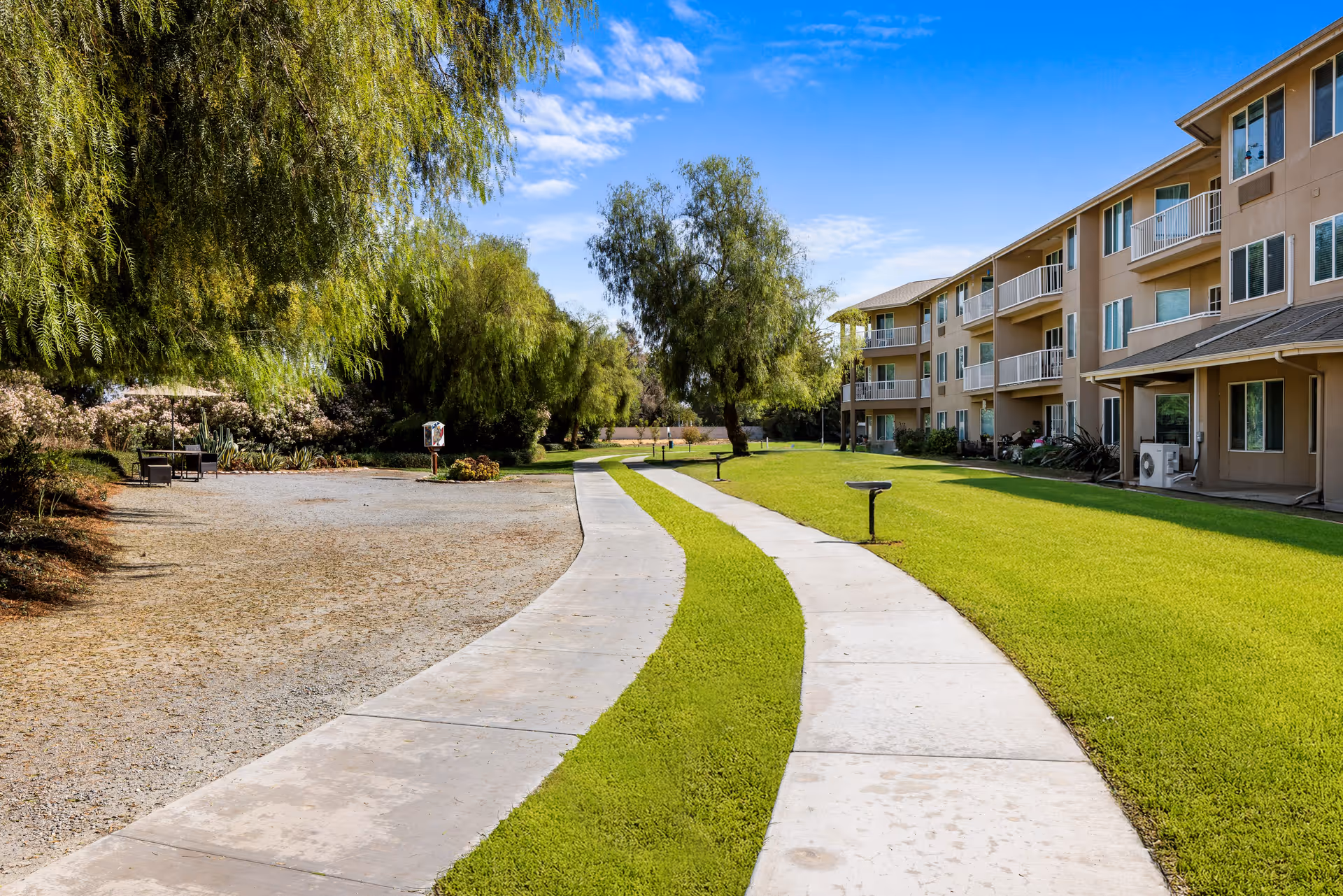 A winding concrete pathway runs through a grassy area with trees and shrubs on one side and a multi-story senior living building with balconies on the other side under a blue sky with some clouds.