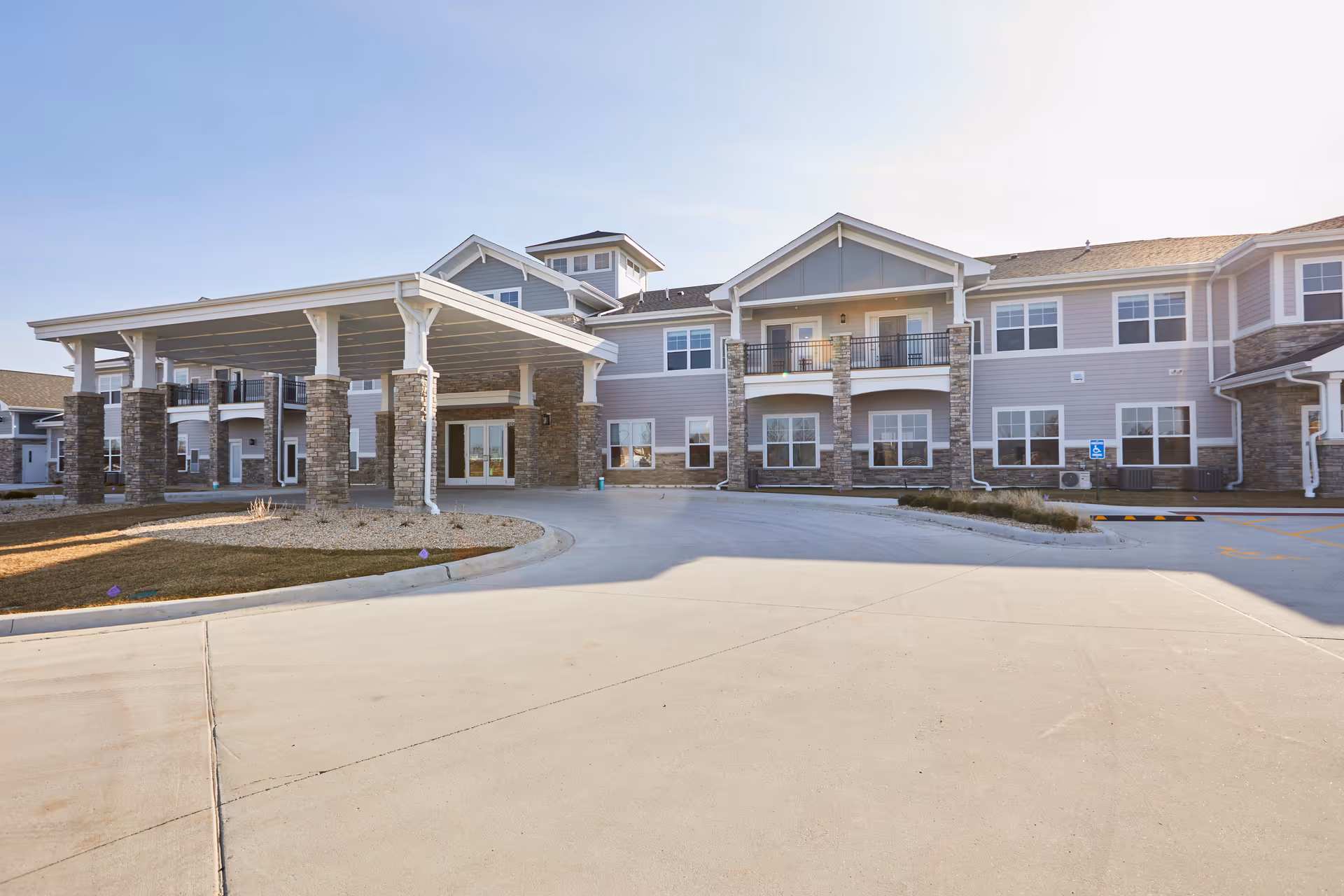 Exterior front view of a senior living facility building with a covered entrance supported by stone pillars, multiple windows, and a driveway in front under a clear sky.