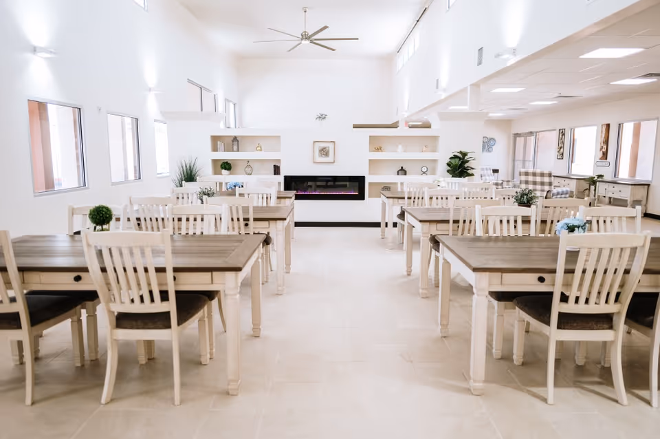 Bright and spacious dining room with multiple wooden tables and white chairs arranged neatly. The room features a modern electric fireplace set into a white wall with built-in shelves decorated with plants and ornaments. Large windows allow natural light to fill the space, and ceiling fans are installed overhead.
